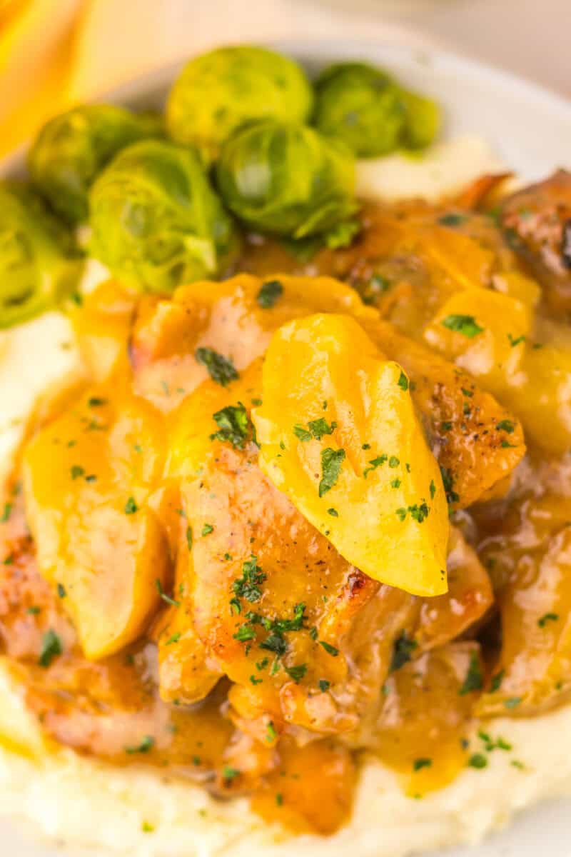 Apple cider chicken with sliced apples and sauce served over mashed potatoes, garnished with chopped herbs, with Brussels sprouts on the side.