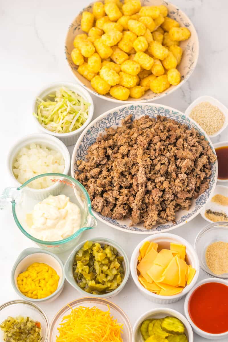 A variety of ingredients in bowls, including tater tots, cooked ground beef, shredded lettuce, sliced cheese, mayonnaise, onions, pickles, relish, grated cheese, and seasonings, arranged on a table.