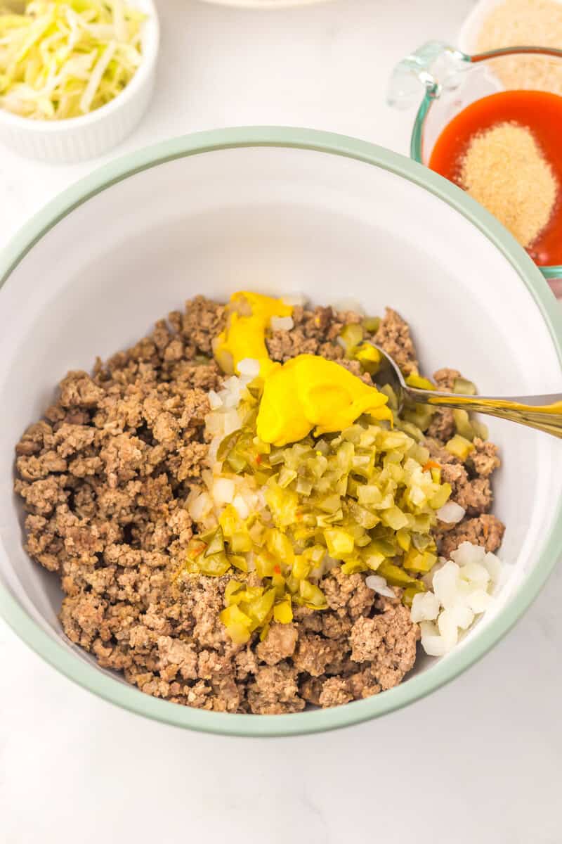 A bowl containing cooked ground beef, chopped onions, diced pickles, and yellow mustard, with a spoon resting inside. Other ingredients are visible in small bowls nearby.