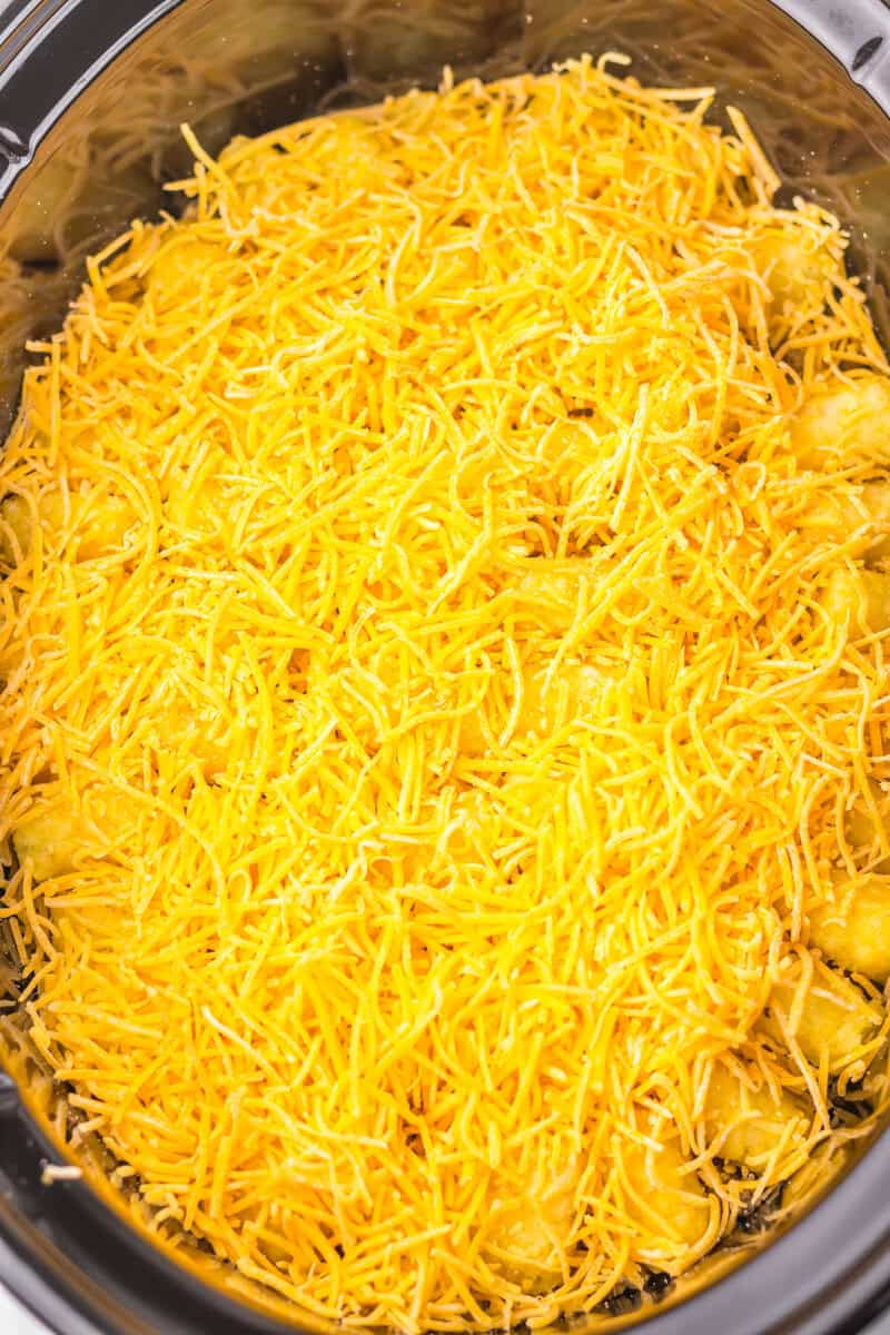 Shredded cheddar cheese covers a layer of tater tots in a slow cooker, ready to be cooked.