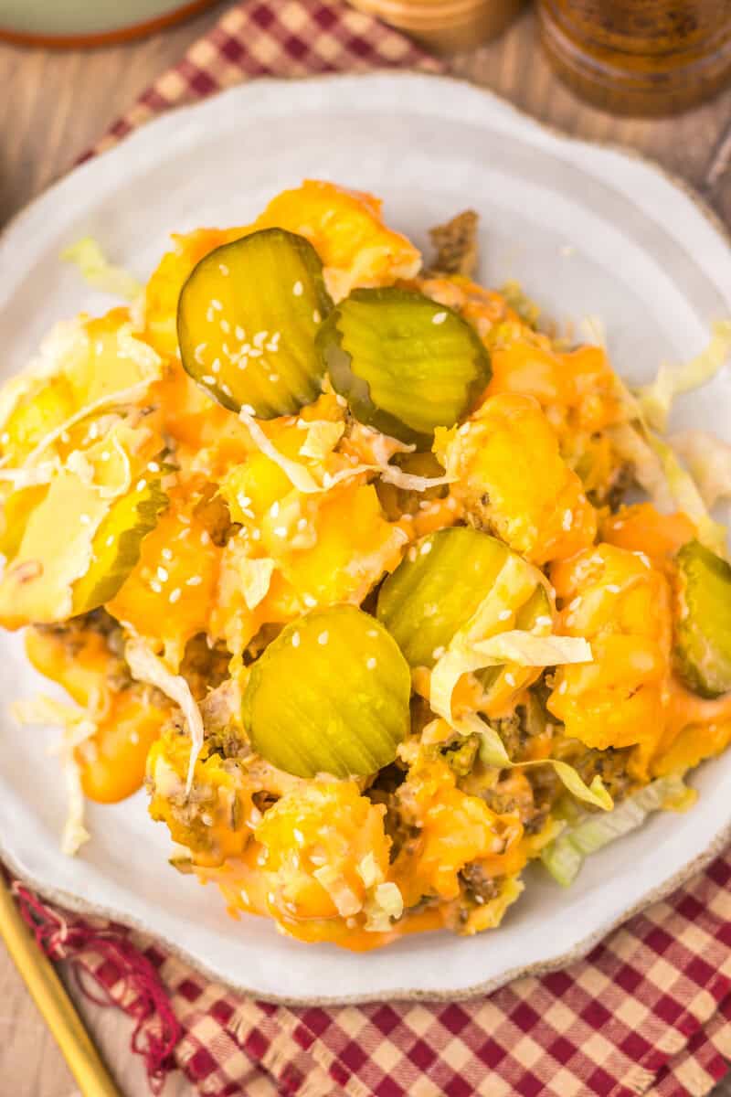 A plate of cheeseburger casserole topped with sliced pickles, shredded lettuce, and melted cheese, served on a white dish over a red plaid napkin.