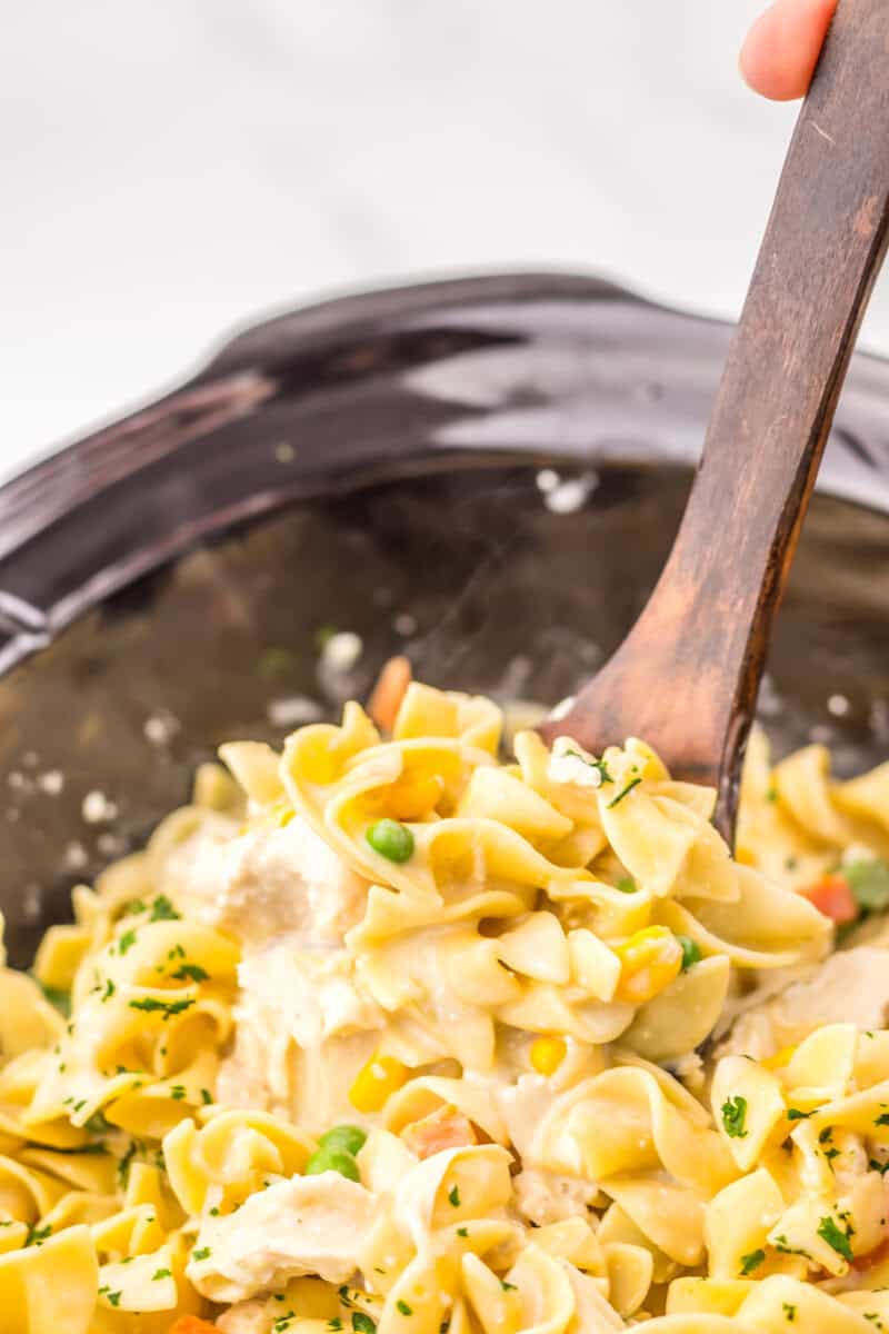 A wooden spoon scoops creamy chicken and noodle casserole with vegetables from a black slow cooker.