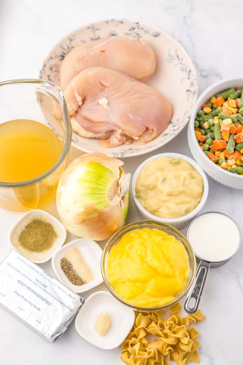 Raw chicken breasts, mixed vegetables, chicken broth, an onion, cream cheese, seasonings, egg noodles, cream of chicken soup, milk, and garlic arranged on a white surface.