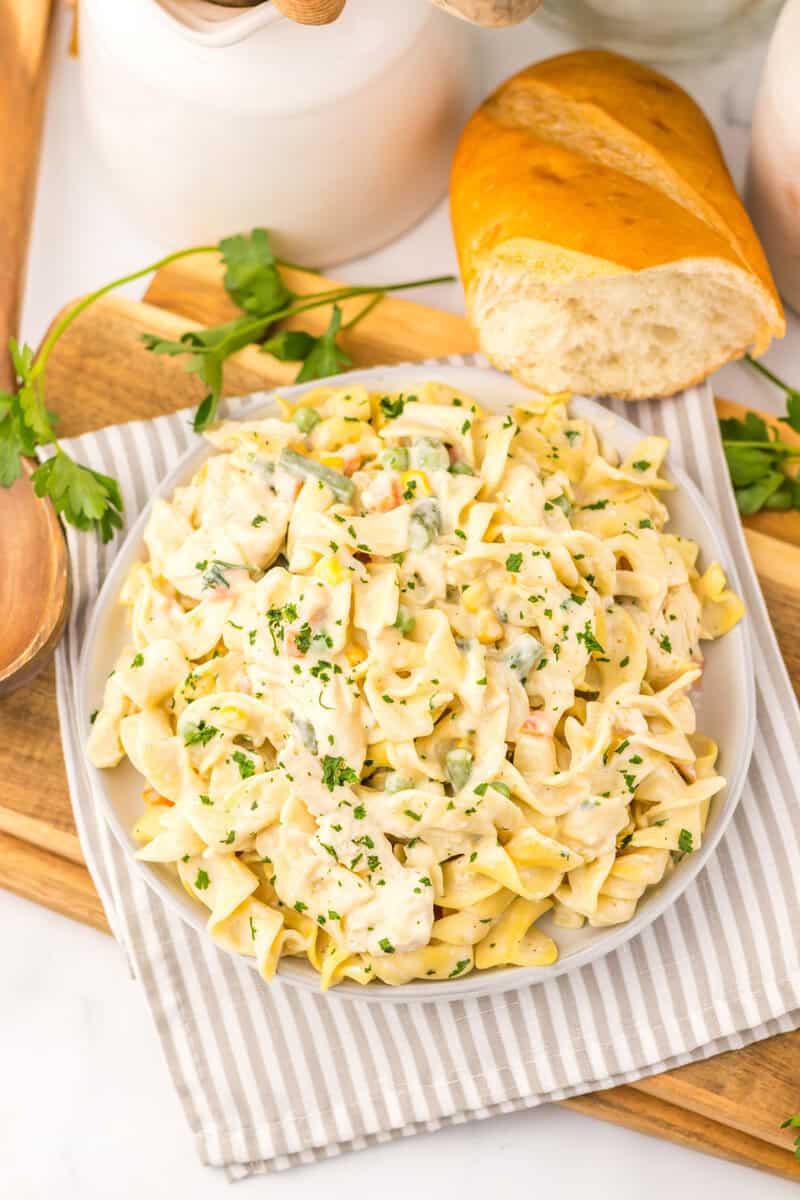 A plate of creamy chicken noodle pasta topped with chopped parsley is served on a striped napkin, with a loaf of bread and fresh parsley beside it.