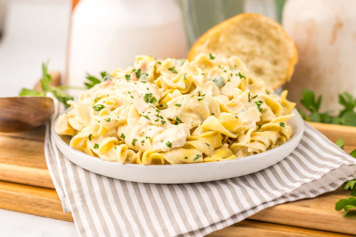 A plate of creamy pasta garnished with chopped herbs sits on a striped napkin, with a slice of bread in the background.