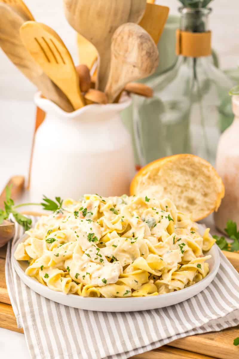 A plate of creamy pasta garnished with parsley sits on a striped cloth, with a slice of bread and kitchen utensils in the background.