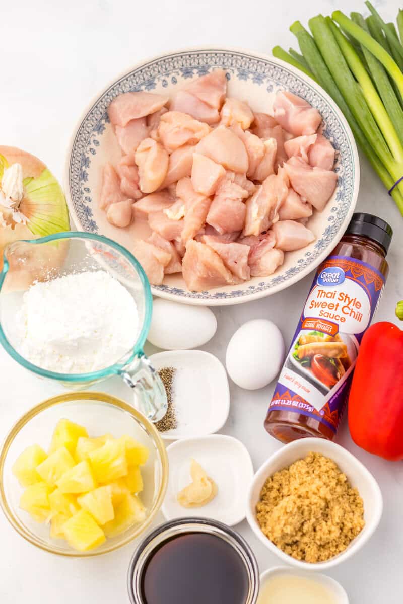Raw chicken, a bottle of thai style sweet chili sauce, eggs, cornstarch, onion, spices, pineapple, brown sugar, and soy sauce on a white background.