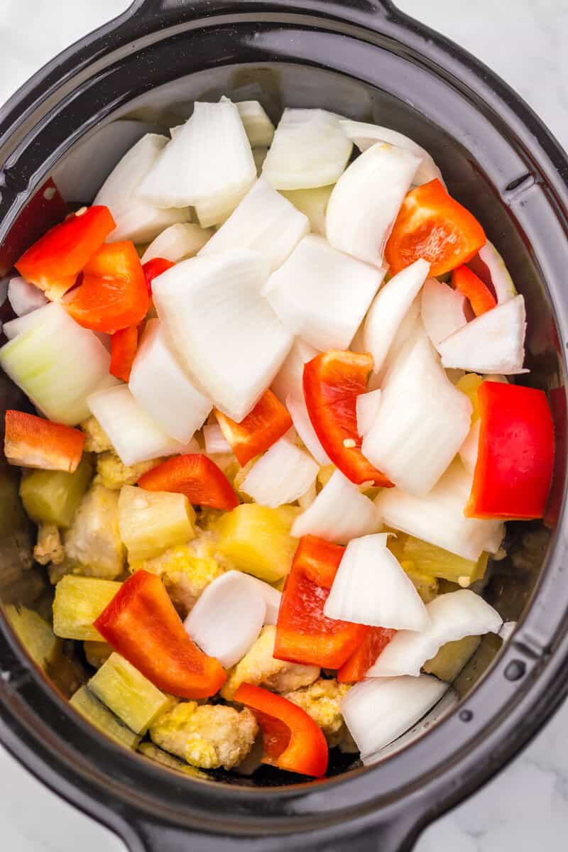 A slow cooker filled with chicken, pineapple, onion, and red peppers.