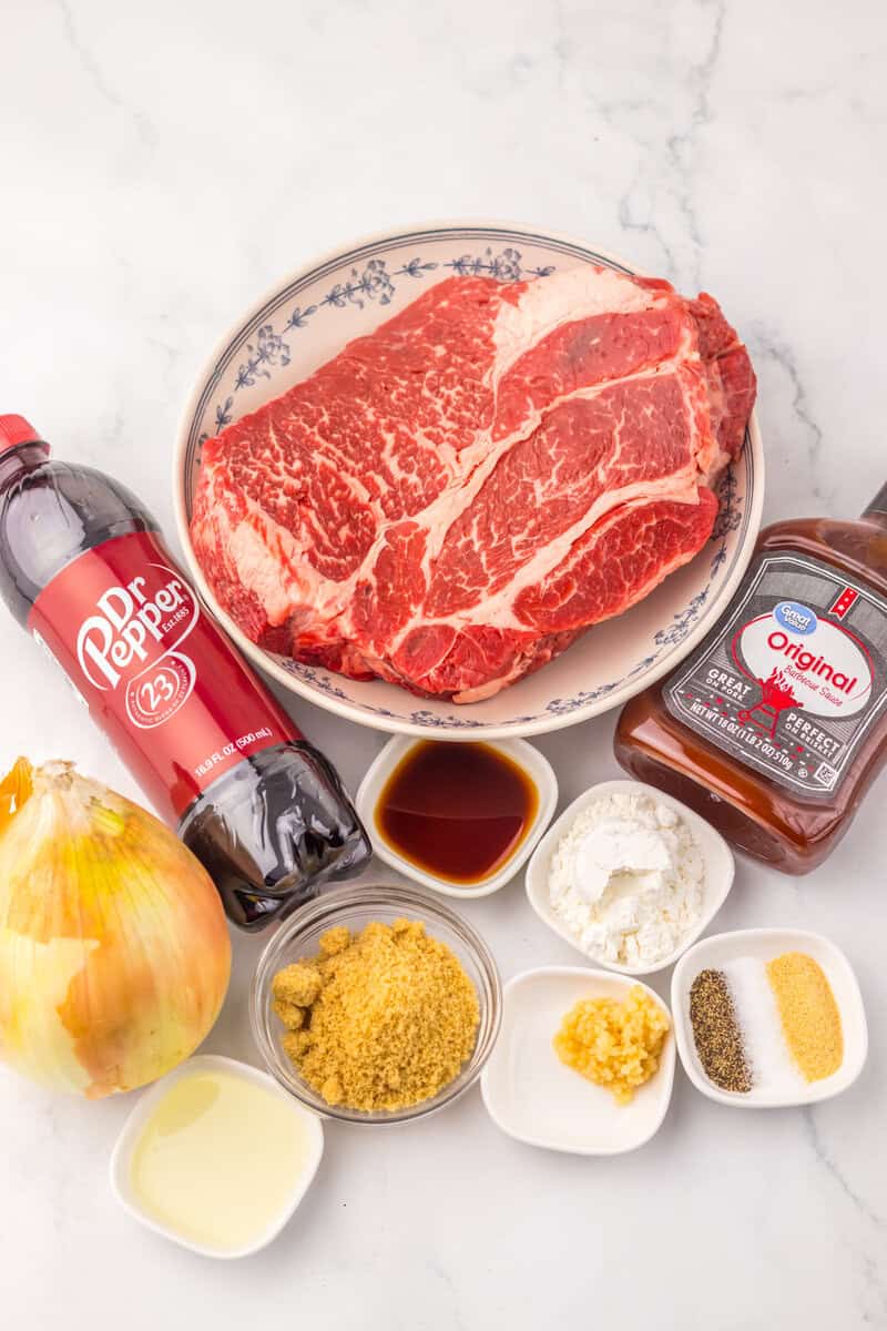 A chuck roast on a white plate surrounded by spices, a bottle of BBQ sauce, and a bottle of Dr. Pepper.