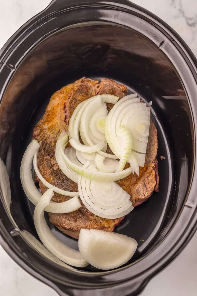 A slow cooker with a chuck roast and sliced raw onions.