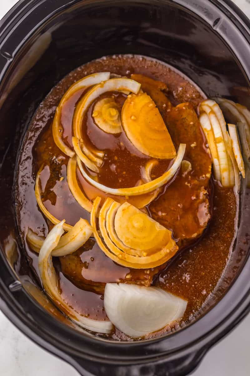 A large piece of beef topped with onions and a brown sauce in a slow cooker.