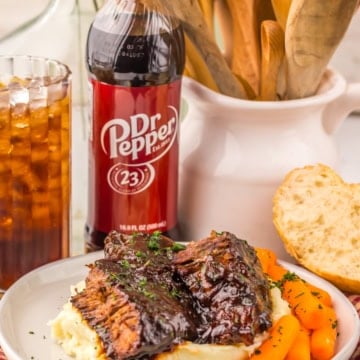 Slow cooker pot roast over mashed potatoes on a plate with carrots. A bottle of Dr. Pepper and a glass of soda are seen in the background.