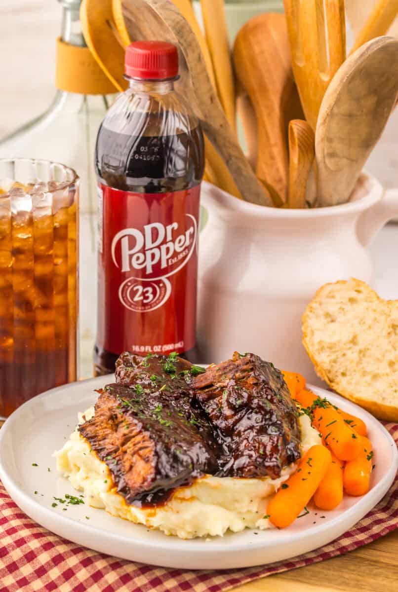 Slow cooker pot roast over mashed potatoes on a plate with carrots. A bottle of Dr. Pepper and a glass of soda are seen in the background.