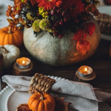 A rustic autumn table setting with plates, candles, small pumpkins, and a floral arrangement in a pumpkin vase.