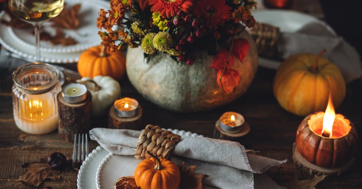 A rustic autumn table setting with plates, candles, small pumpkins, and a floral arrangement in a pumpkin vase.