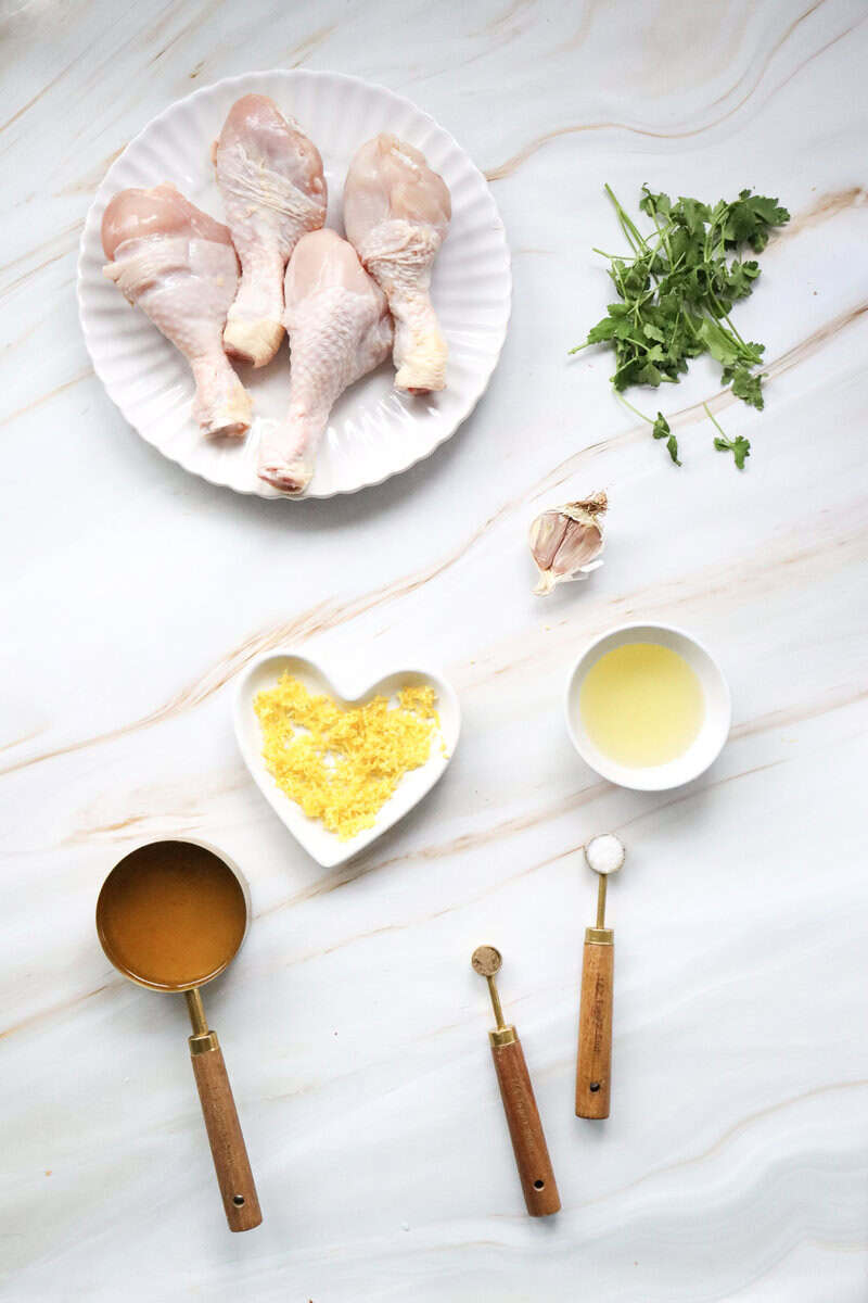 A marble countrer top with a white plate of raw chicken drumsticks, fresh parsley, small bowls of lemon juice and lemon zest, part of a garlic head, and measuring cups filled with salt, pepper, and chicken broth.