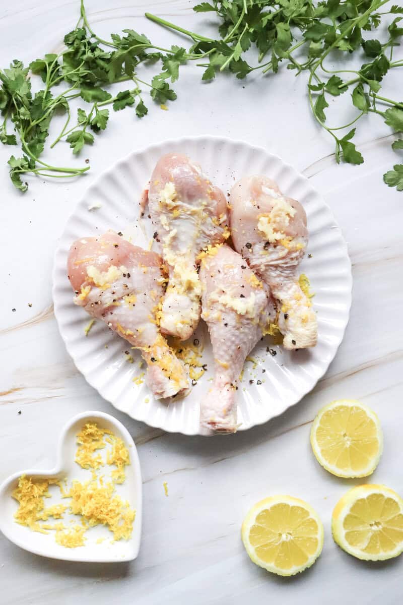 A white plate with chicken drumsticks covered in a mixture of lemon zest, lemon juice, salt, pepper, and garlic.