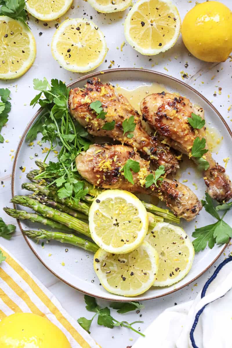 Air fryer drumsticks on a white plate with asparagus and lemon slices, topped with parsley.