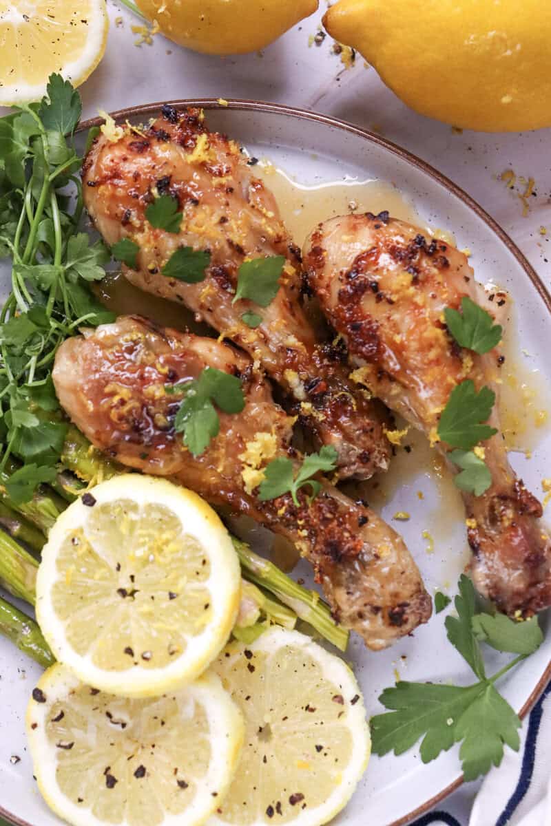A plate of air fryer chicken drumsticks with lemon garlic seasoning topped with parsley, seen on a white plate with asparagus and lemon slices.