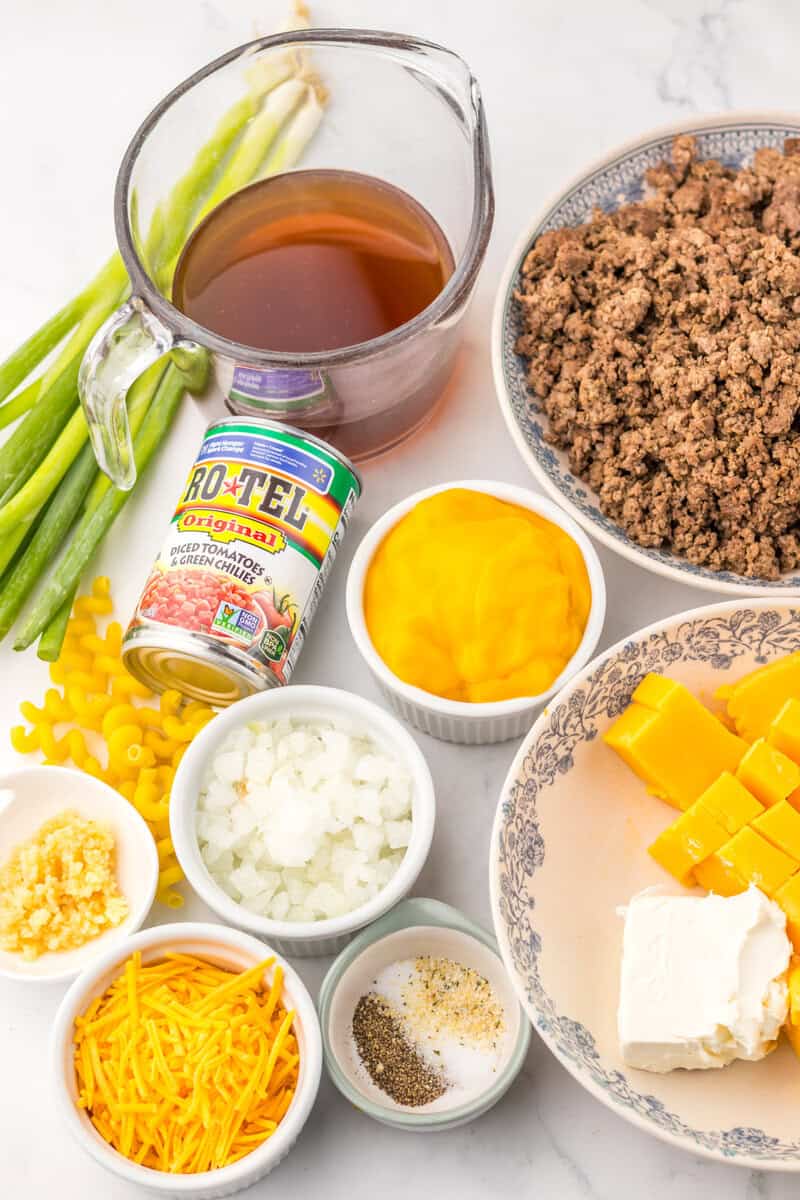 Individual bowls of ground beef, cheeses, diced onions, garlic, and spices. A mesuring cup of beef broth, green onions, dry pasta, and a can of Rotel can also be seen.