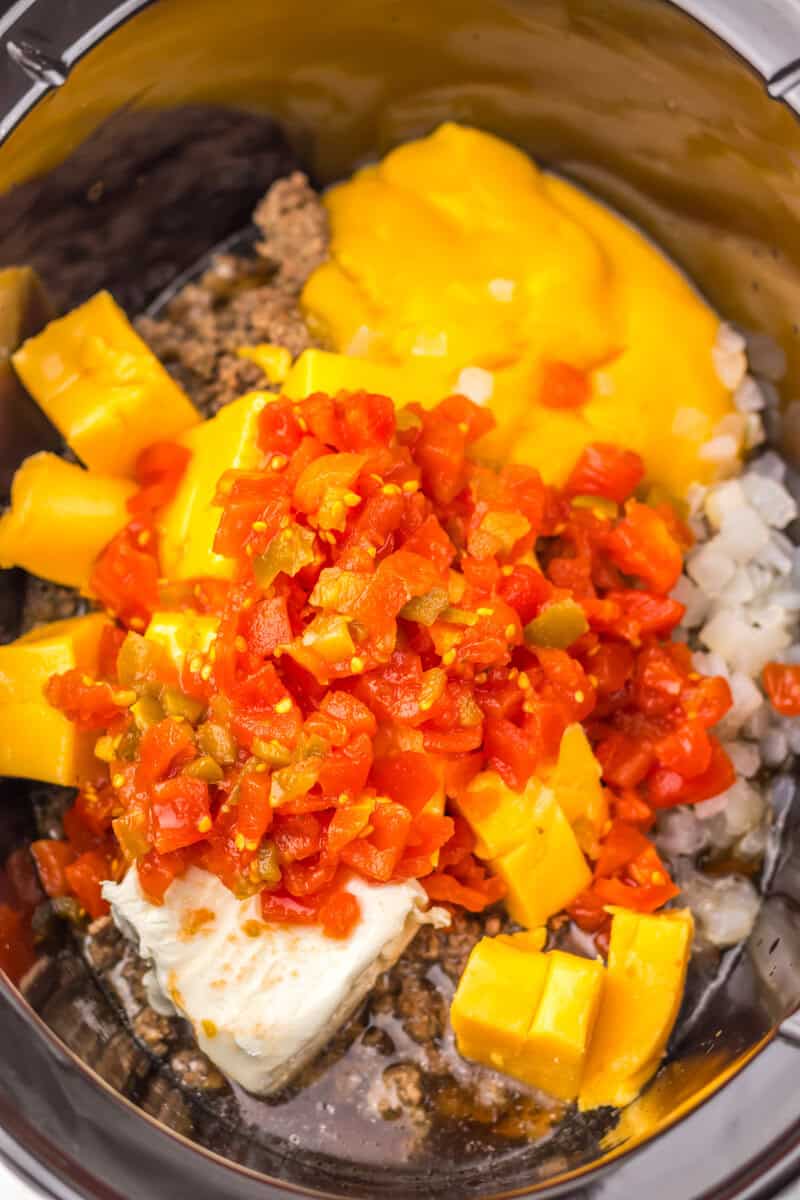A slow cooker with Rotel, cubed Velveeta, cream cheese, ground beef, and diced onions.
