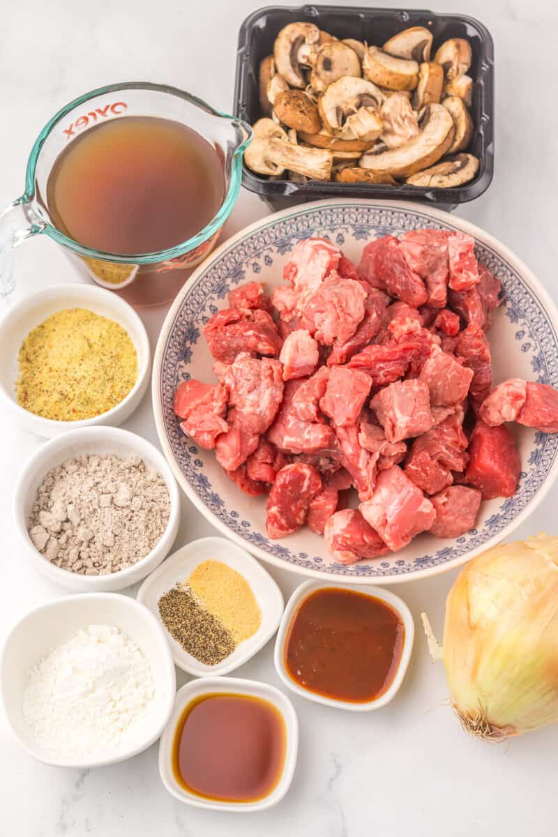 A blue and white bowl of raw beef with individual bowls of seasonings and sauces, an onion, mushrooms, and brother on a white counter.