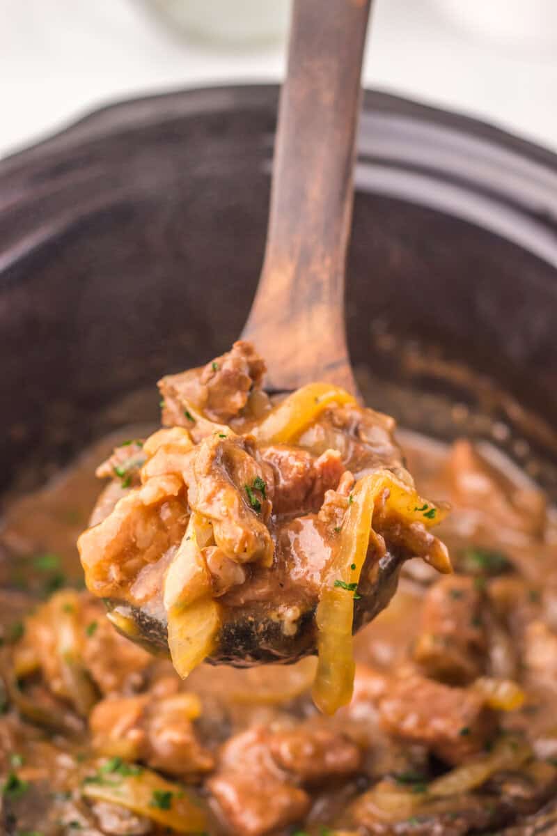 A wooden spoon lifting Texas Roadhouse style steak tips out of a slow cooker.