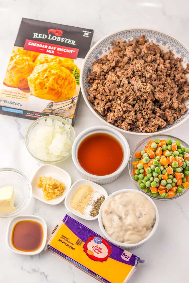 Bowls of peas and carrots, ground beef, spices, sauces, cream of mushroom soup, a box of Red Lobster Cheddar Bay Biscuit Mix, and a block of sharp cheddar cheese on a white surface.