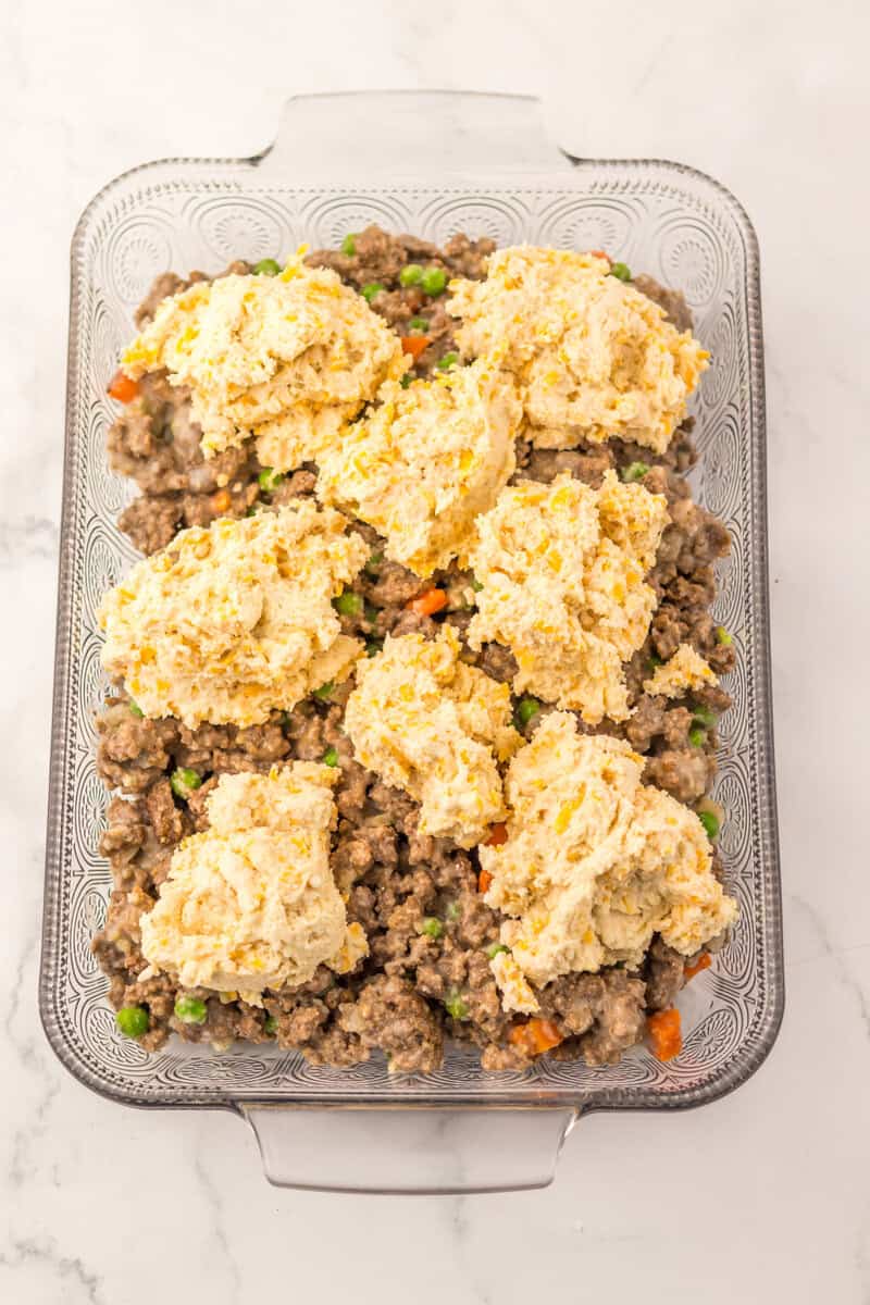 Uncooked biscuit dough on top of a beef and vegetable mixture in a clear glass baking dish.