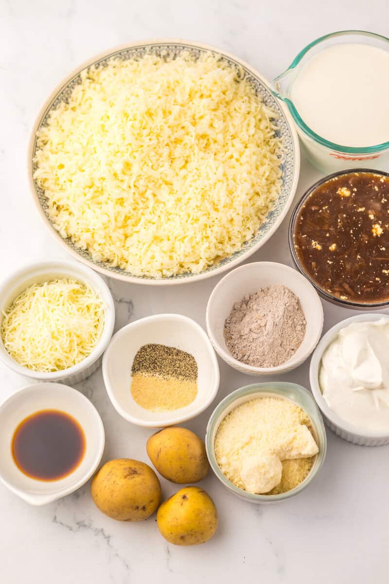 A white countertop with individual bowls holding cheese, sour cream, spices, French onion soup, cream, and potatoes.