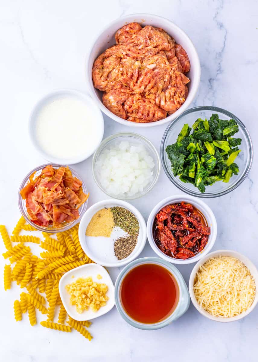 Individual bowls of sausage, kale, sundried tomatoes, broth, cheese, bacon, cream, garlic, onions, and spices on a white surface with dried rotini pasta.