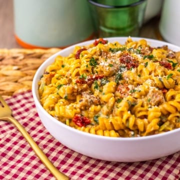 A white bowl of creamy Tuscan pasta on a table with a red and white checkered napkin.