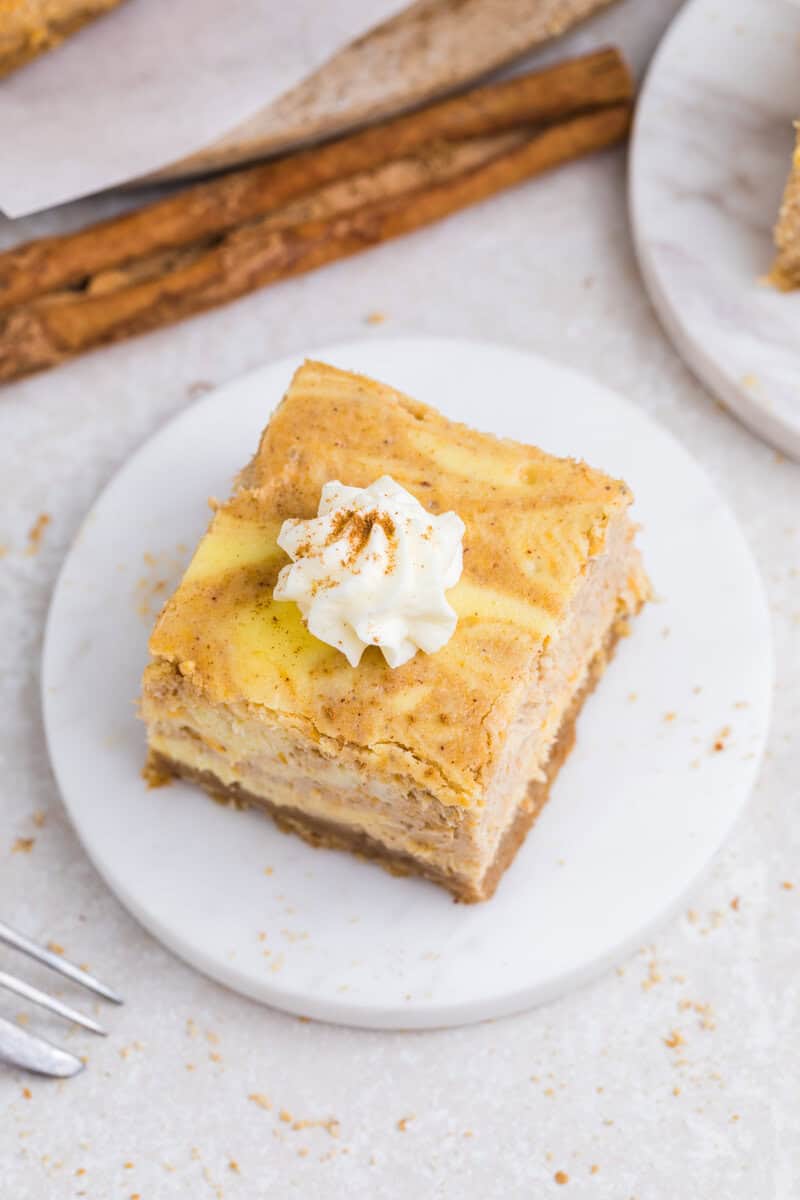 A single pumpkin cheesecake bar on a white plate topped with whipped cream and cinnamon.