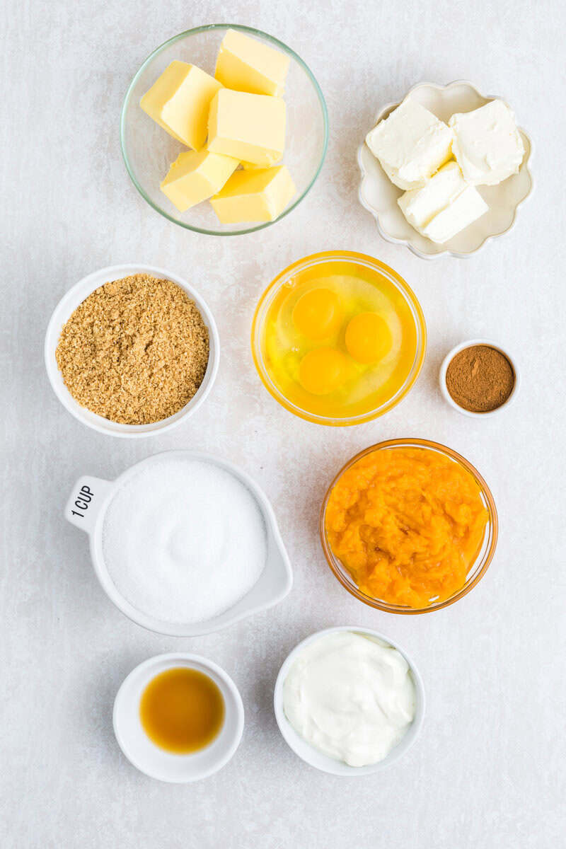 Individual bowls of cream cheese, butter, eggs, pumpkin, spices, graham cracker crumbs, sour cream, sugar, and vanilla on a white surface.