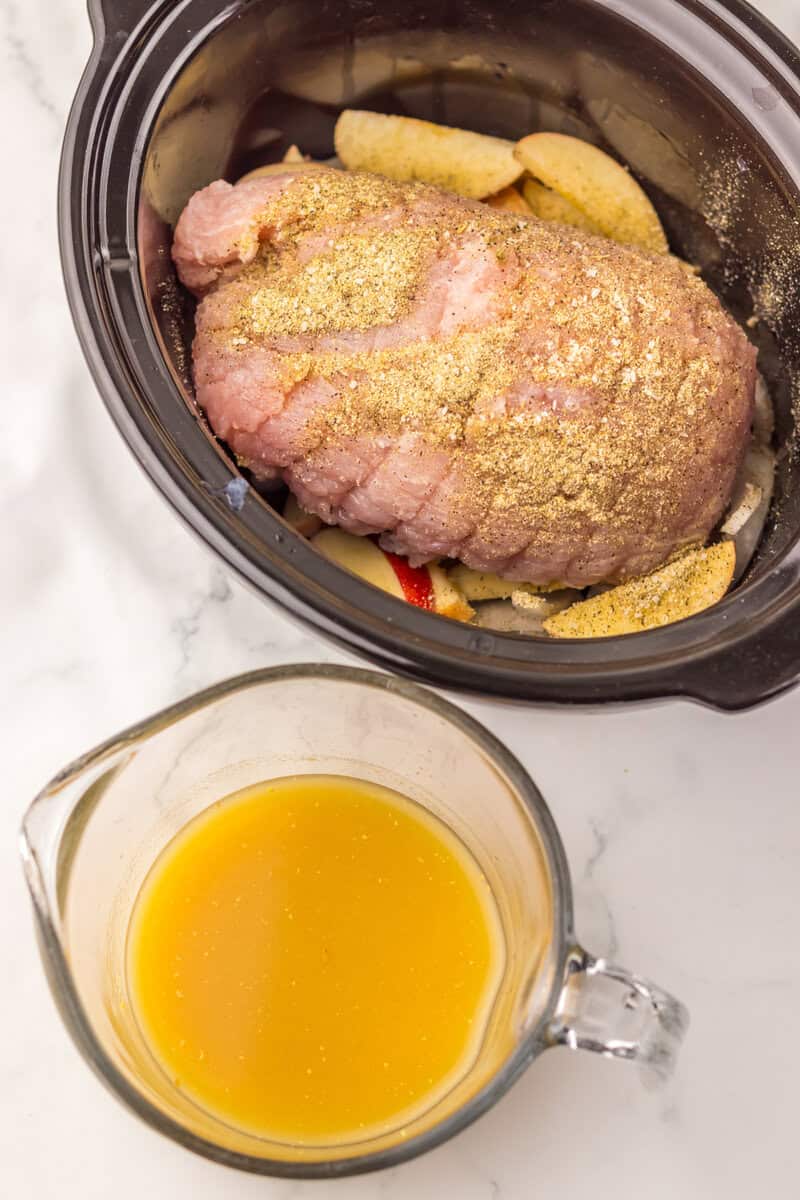 An uncooked turkey breast and apple slices in a slow cooker with a glass mixing cup of a golden liquid nearby.