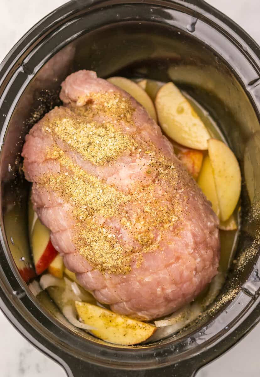An uncooked turkey breast in a slow cooker with apples and onions underneath.