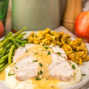 Slow cooker turkey breast with gravy on mashed potatoes, served with green beans and stuffing on a white plate.