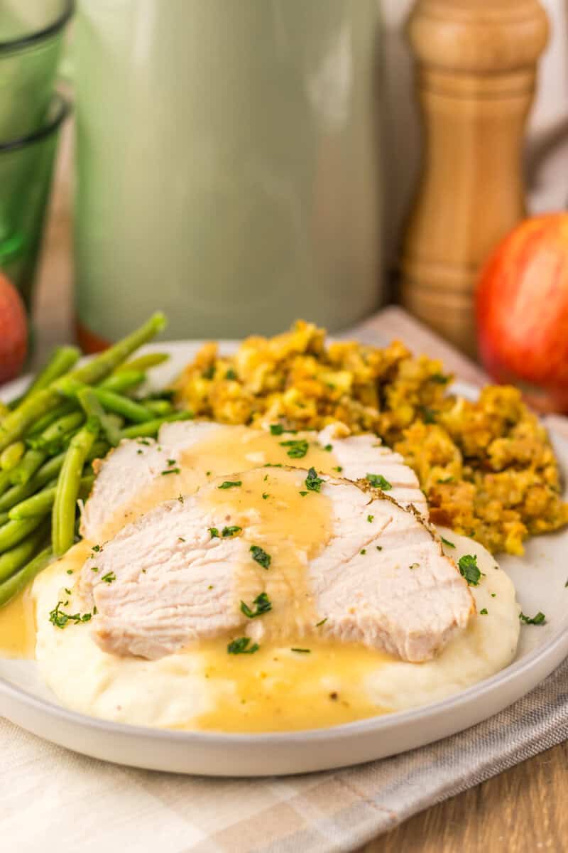 A plate of slow cooker turkey breast, sliced and topped with gravy, alongside green beans and stuffing. Apples, a pitcher, and a pepper grinder re seen in the background. 