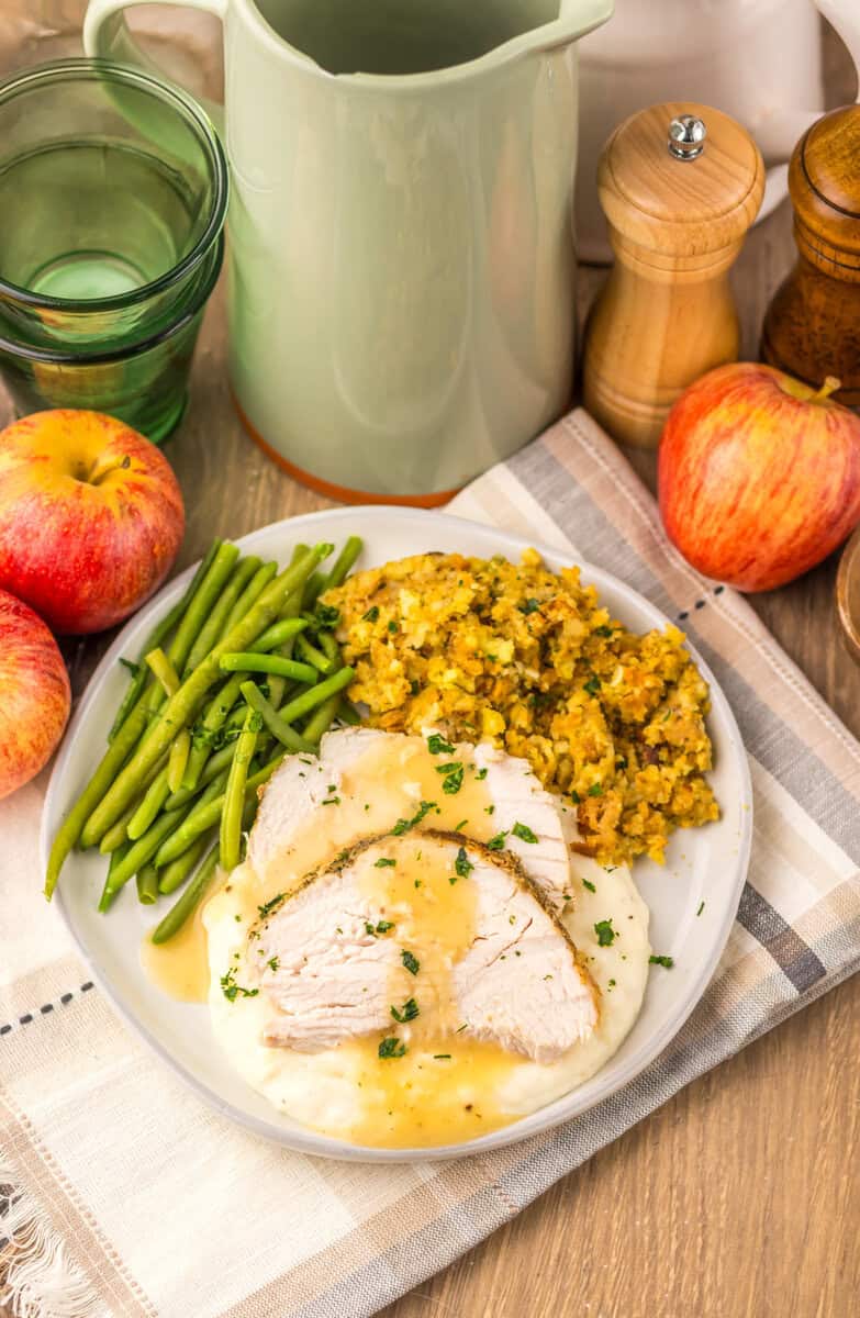 A plate of sliced turkey, green beans, and dressing surrounded by red apples and kitchen utensils.
