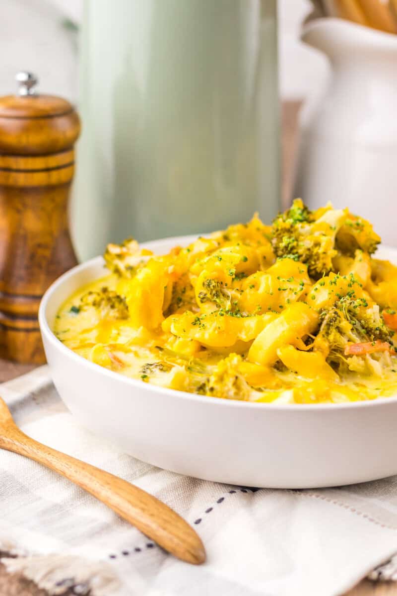 A white bowl of broccoli cheddar tortellini soup with a wooden pepper grinder and a light green pitcher in the background.