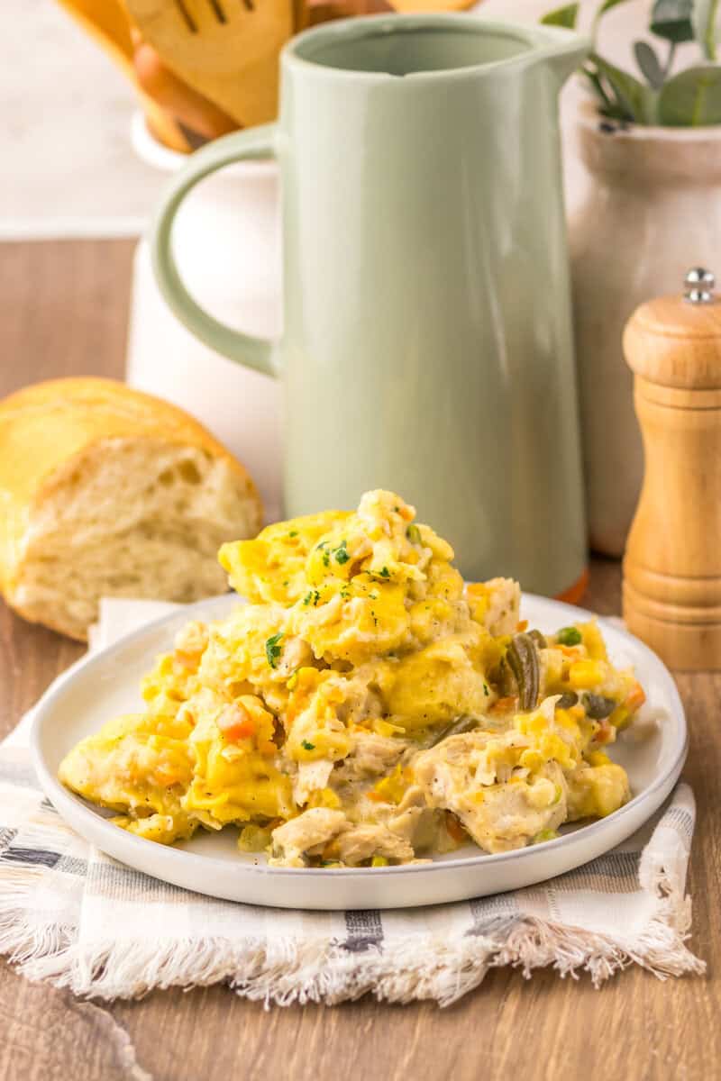 A serving of Crock Pot chicken and dumplings on a white plate with a pitcher, a pepper grinder, and a piece of French bread in the background.
