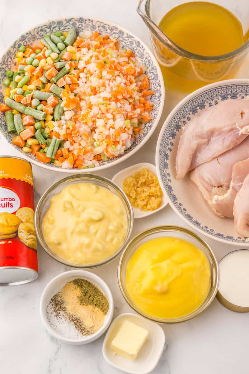 A bowl of frozen vegetables, raw chicken, broth, condensed soup, garlic, butter, seasoning, and a can of biscuits on a white surface.
