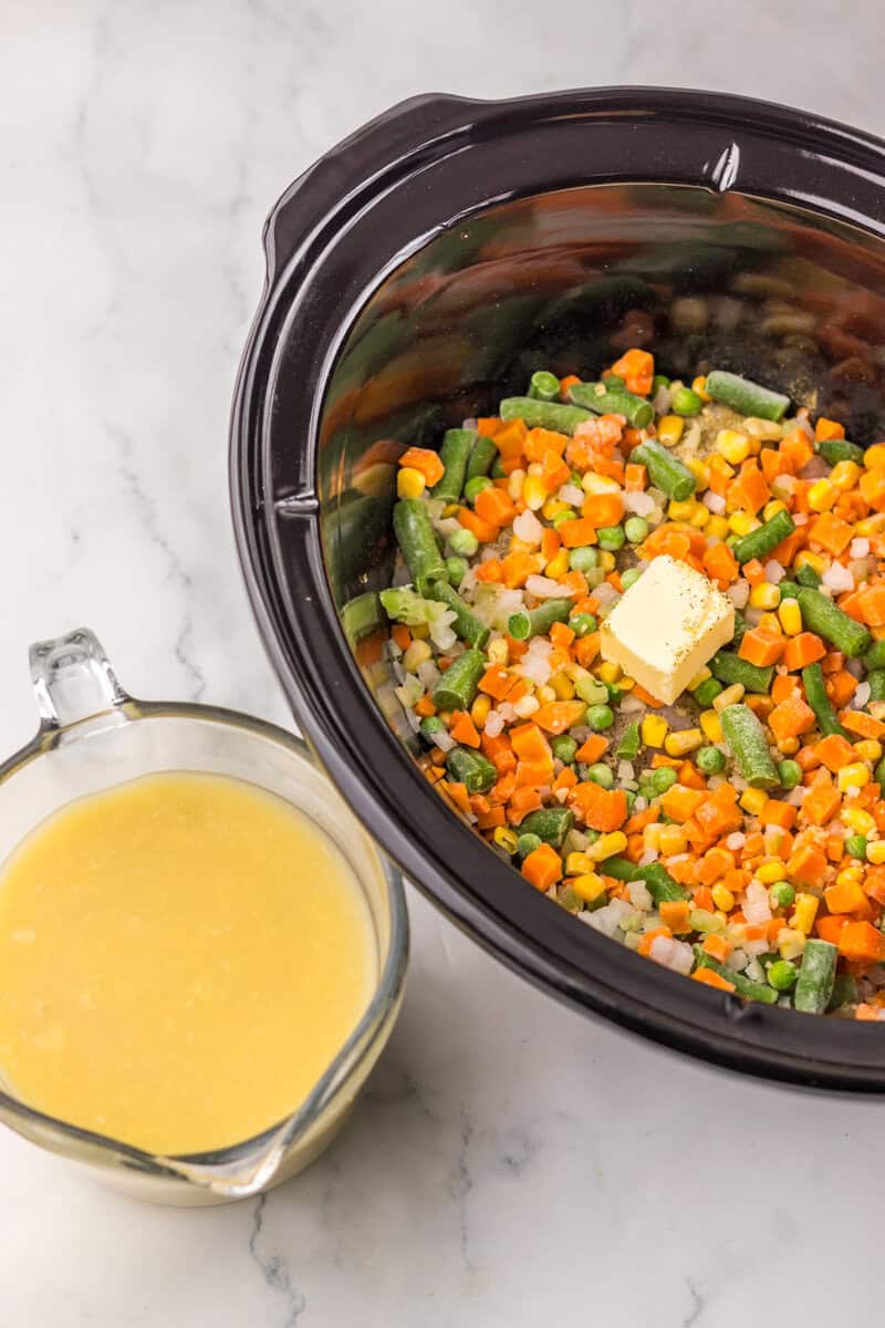 A black Crock Pot filled with frozen vegetables and butter with a glass measuring cup of condensed soup nearby.