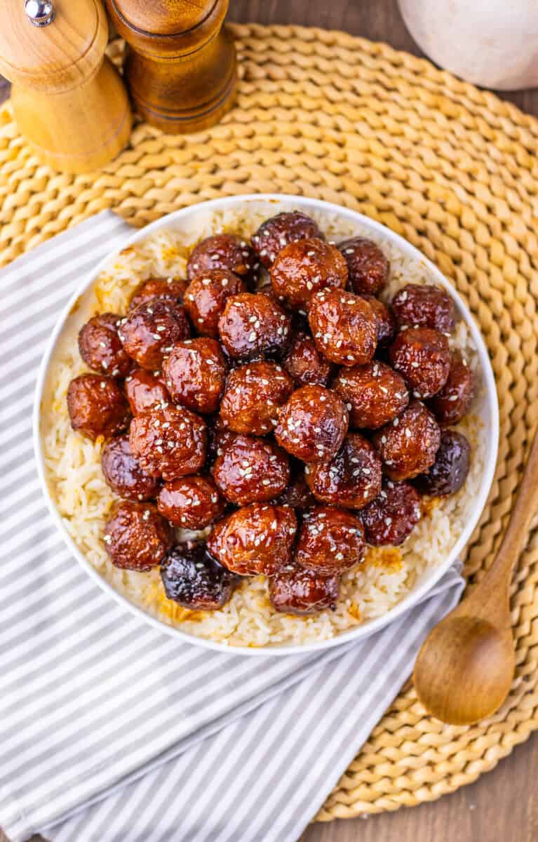 A bowl of sticky sesame meatballs on top of brown rice on a table.