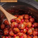 Slow cooker filled with cranberry slow cooker meatballs in sauce, garnished with fresh cranberries and herbs, with a wooden spoon for serving.