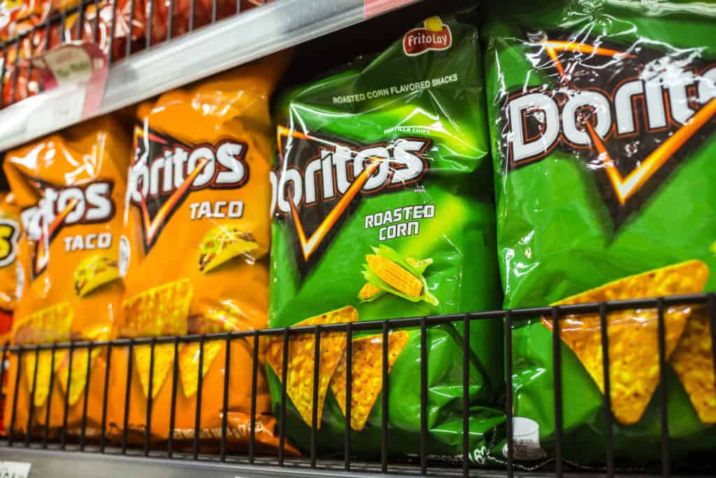 Bags of Doritos in Taco and Roasted Corn flavors are displayed on a store shelf behind a wire rack, inviting customers to wonder,