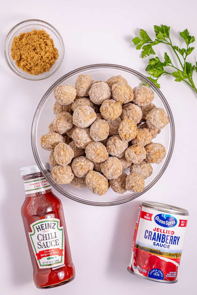 Individual bowls of meatballs and brown sugar with a can of cranberry sauce and a jar of Heinz chili sauce on a white surface.