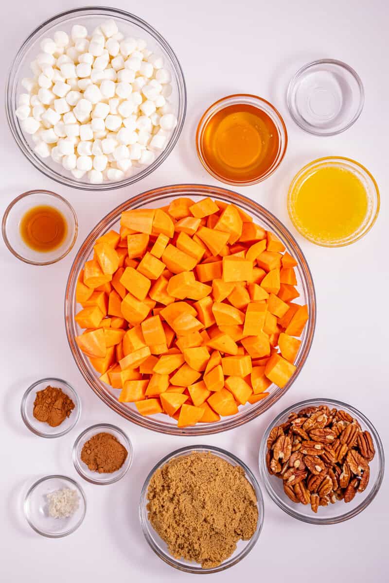 Individual bowls of sweet potatoes, marshmallows, pecans, maple syrup, and other ingredients on a light surface.