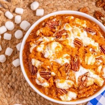 A bowl of mashed sweet potatoes topped with marshmallows and pecans on a tablecloth.