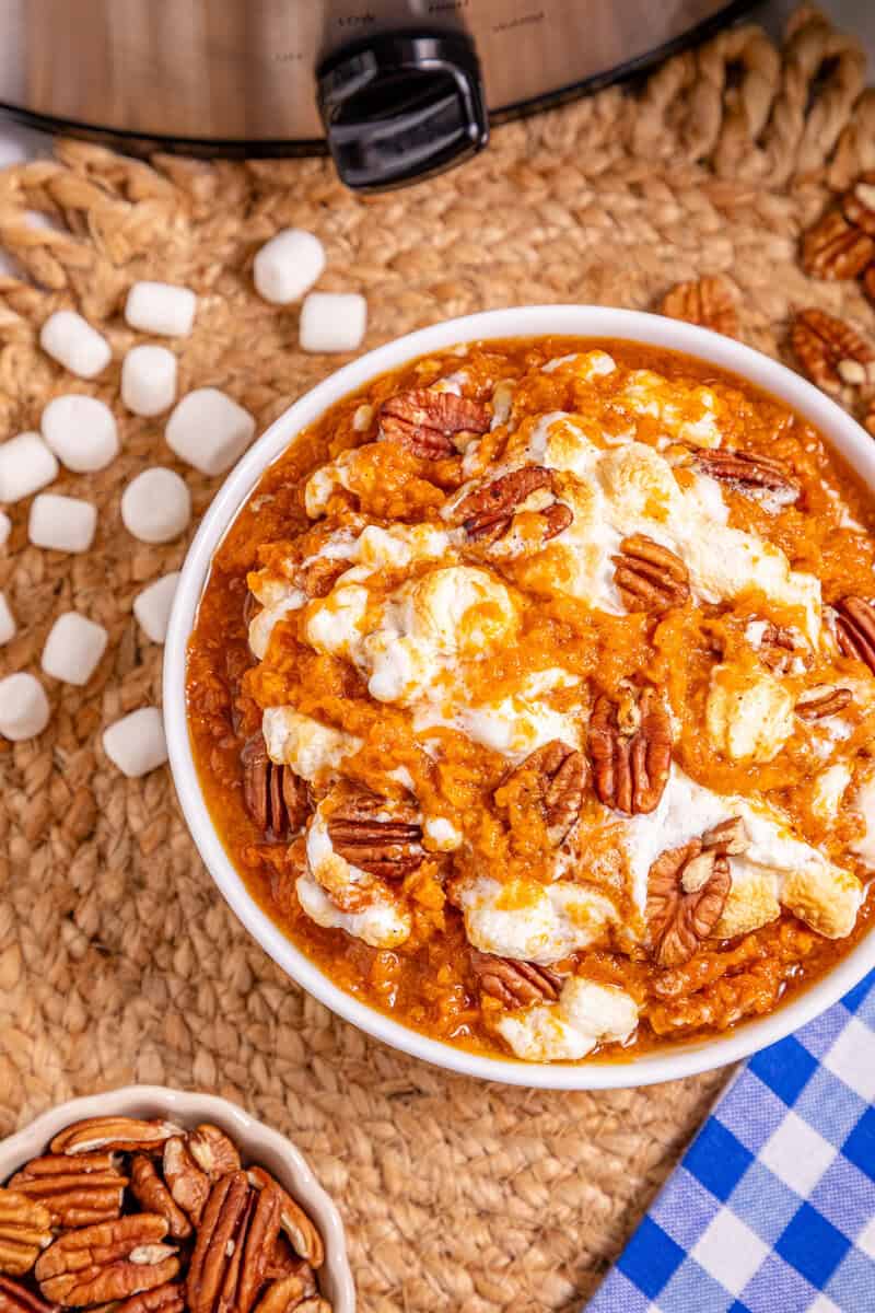 A bowl of mashed sweet potatoes topped with marshmallows and pecans on a tablecloth.